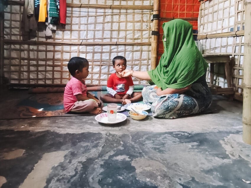 A woman sits on the ground feeding a young child, while another child watches them closely.