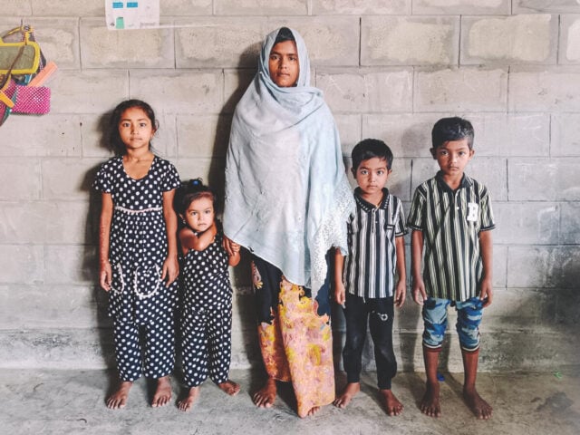 A woman in a headscarf stands with her four children in front of a gray cement block wall. Two wear polka dots, the others wear stripes.