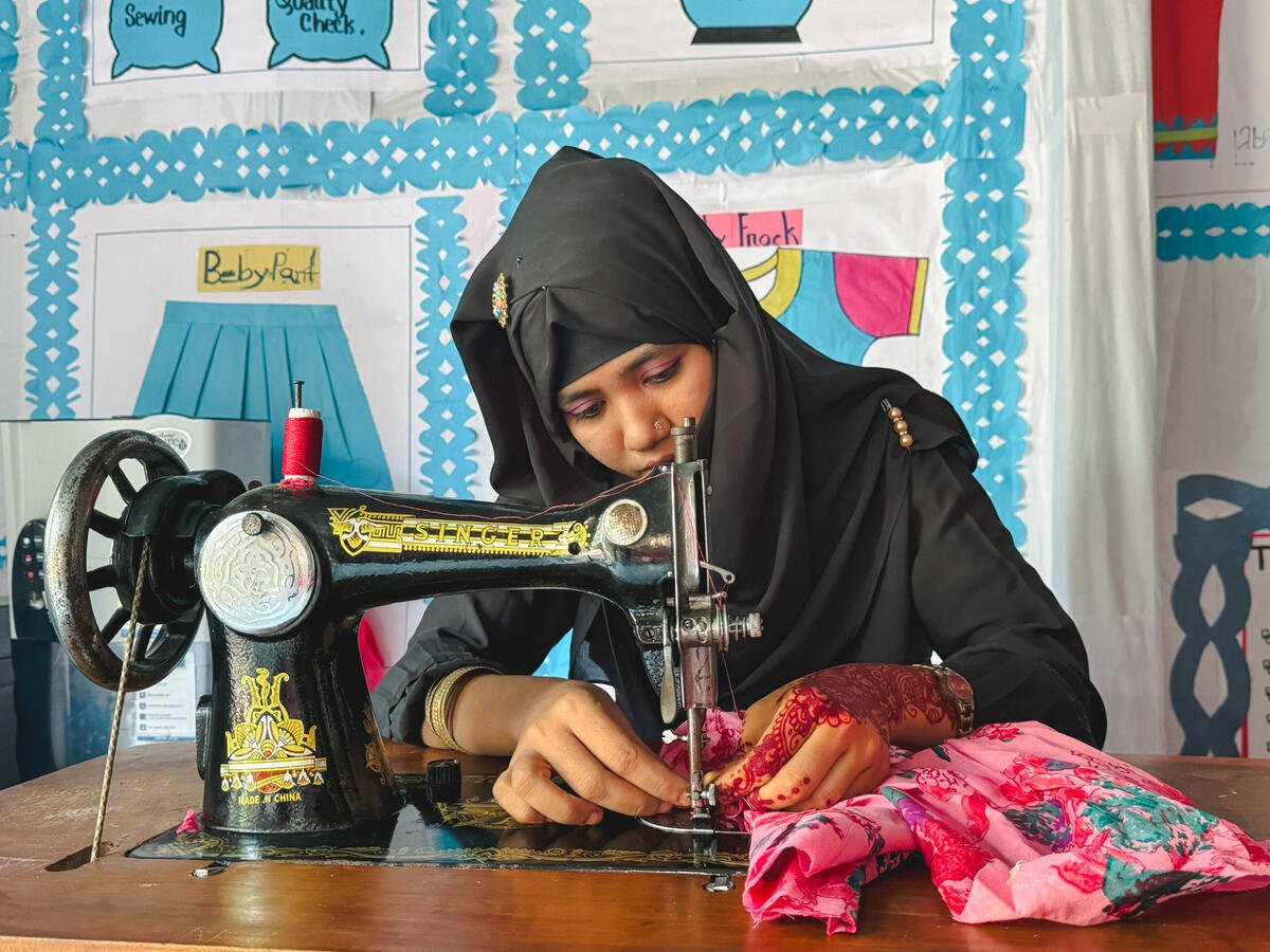 A woman wearing a headscarf uses a sewing machine, guiding pink fabric beneath the needle with both hands.