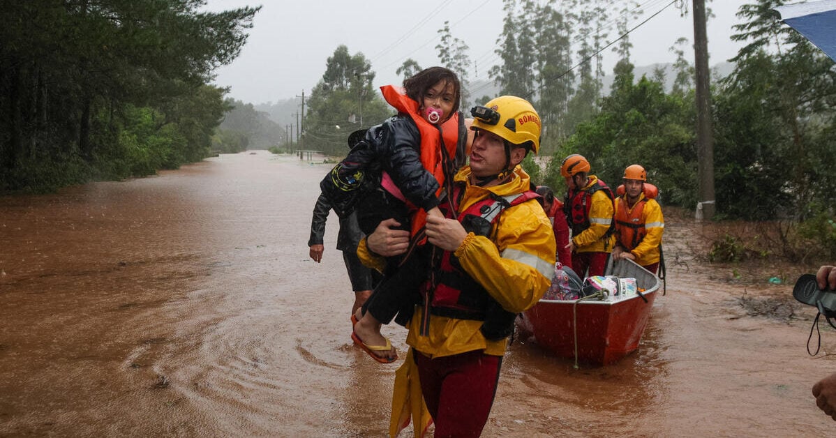 A man in a hard hat carries a young girl with a pacifier and life vest through floodwaters, with a rescue team and boat behind them.