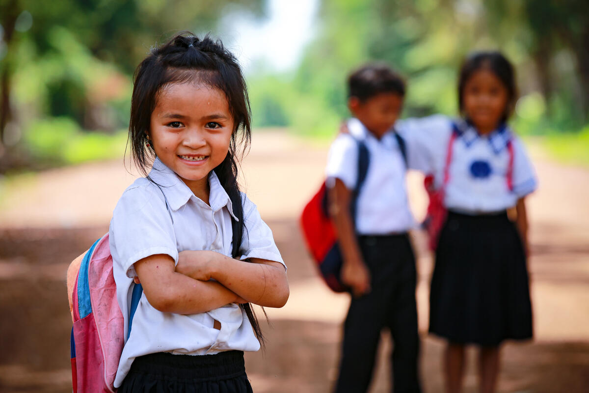 A girl stands with arms crossed in the foreground. Two other children in school uniforms stand in the background.