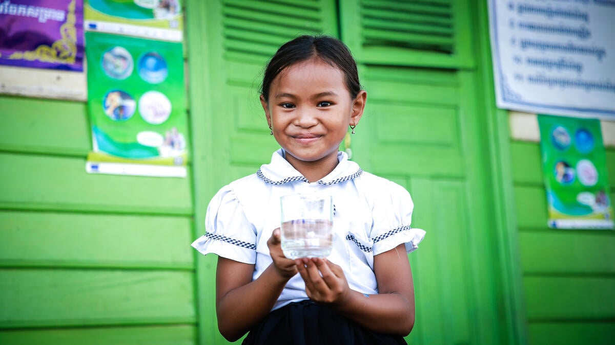 A girl in a white blouse with a blue gingham trim stands before a bright green structure and looks at the camera while holding a glass of clean water. 