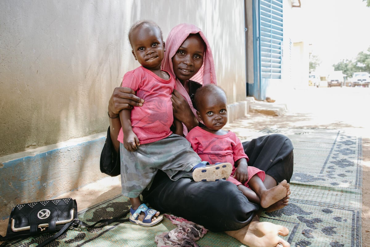 A woman in a pink headscarf sits on the ground on a blue carpet, gazing into the camera as she holds a healthy-looking young boy in her arms. The boy’s twin, wearing a matching pink shirt, sits on her lap.