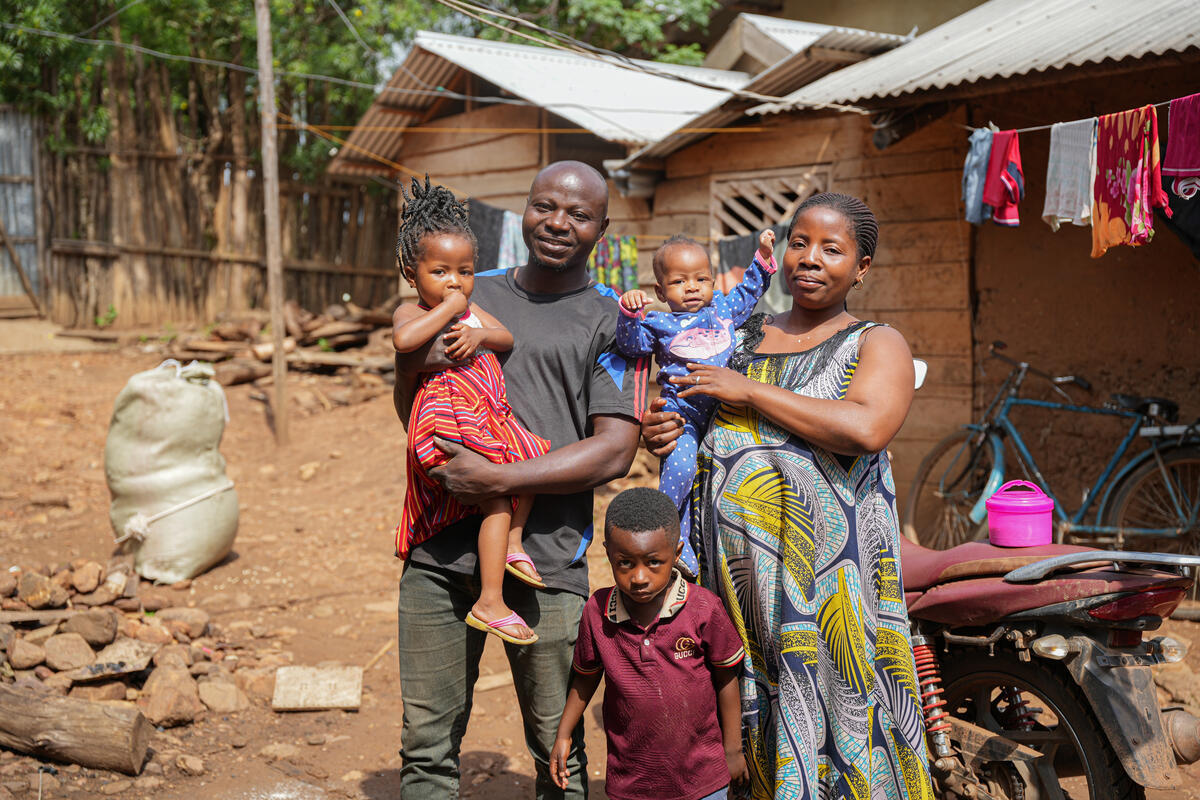 A family of five stands outside their home, with the parents holding two young children while another child stands in front.