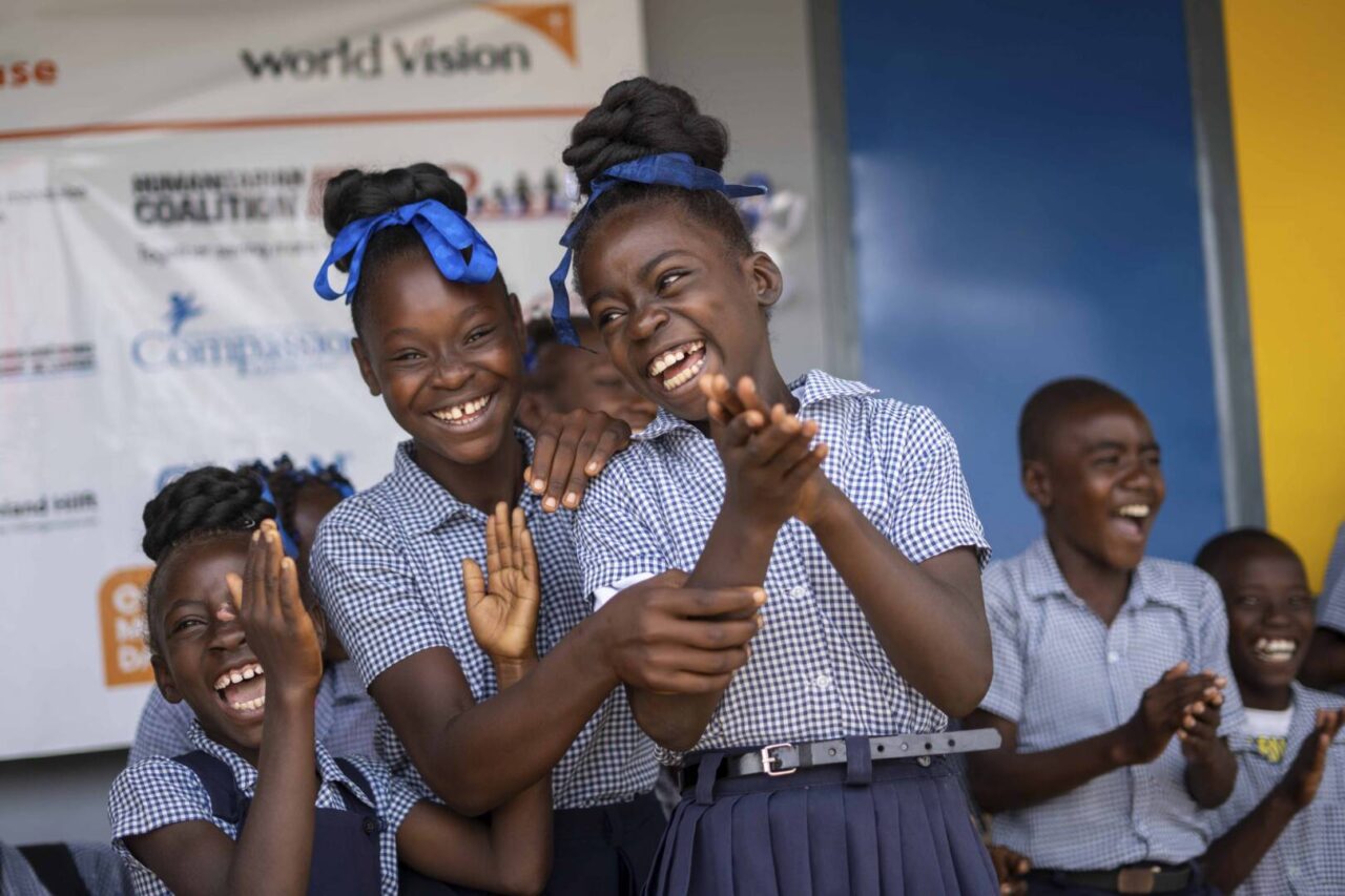 Haitian girls in school uniforms and blue hair bows laugh and clap in joy.