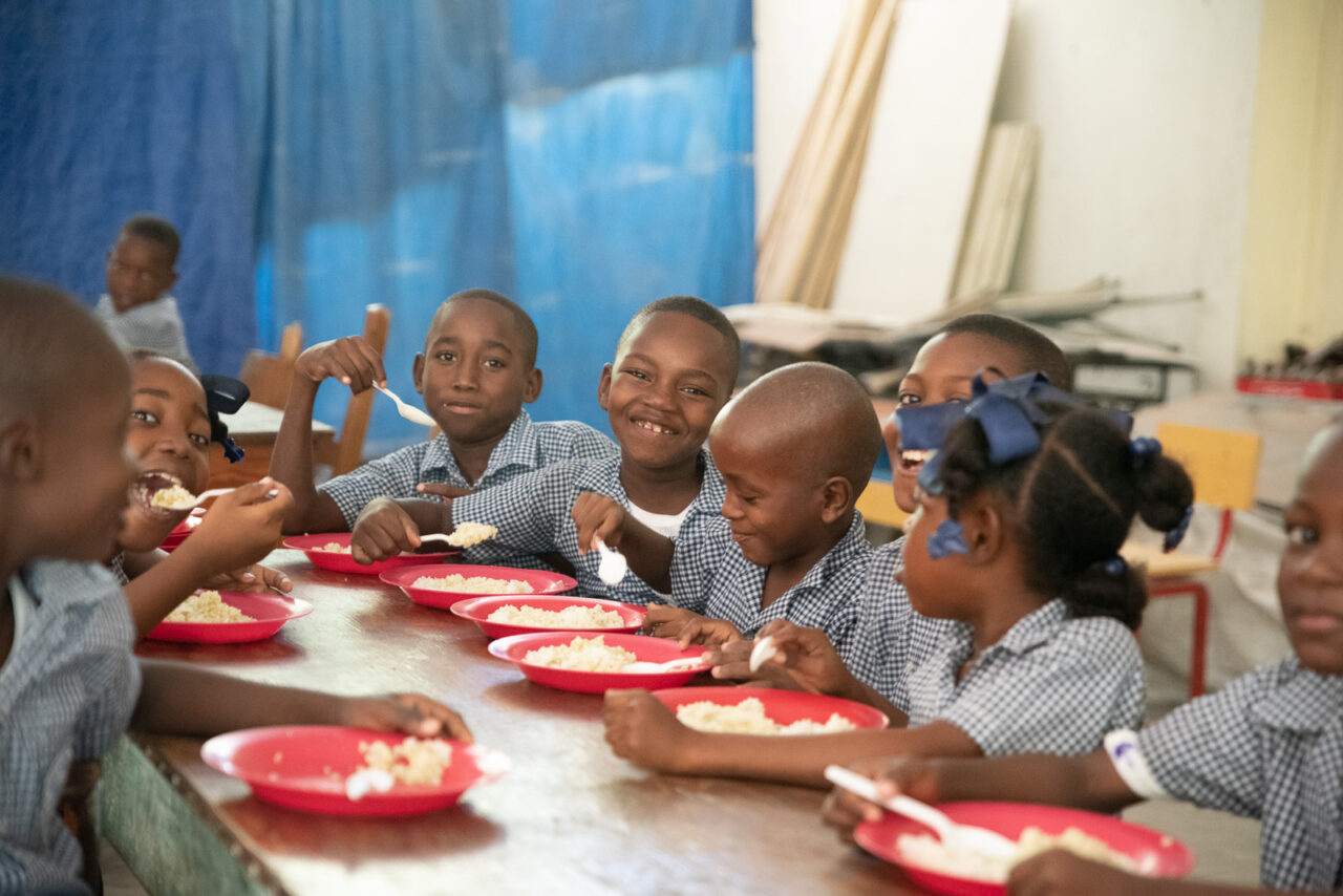 Children wearing navy-and-white gingham school uniforms sit at a lunch table with red plates holding their meals.