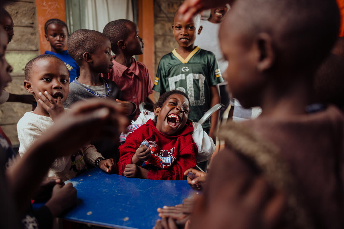 Group of joyful children with an adult, celebrating by clapping, around one child in a wheelchair laughing heartily.