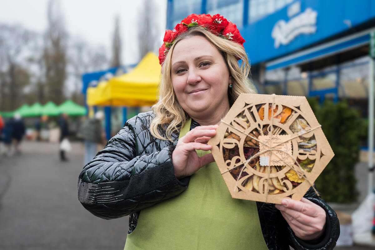 Adorned with a red rose wreath, a blonde woman proudly lifts a hexagon-shaped box filled with dried fruits.