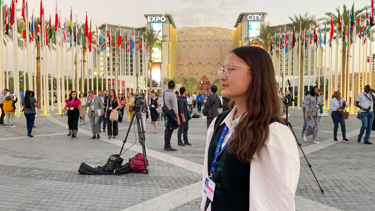 A bespectacled girl, in a white shirt and navy vest, stands in a public square amid flags.