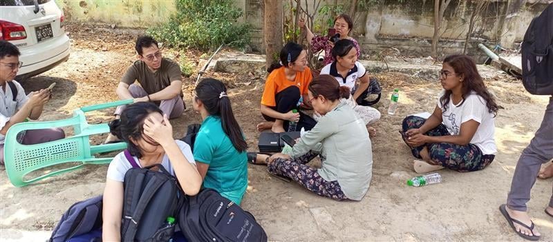 A group of people sit on the ground outside.