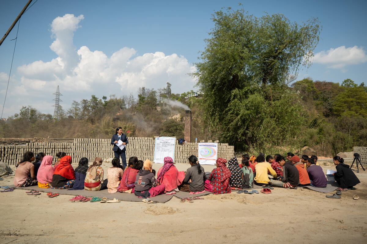 A group of brick workers in Nepal sit on the ground facing a standing person who is speaking under a blue sky with clouds.