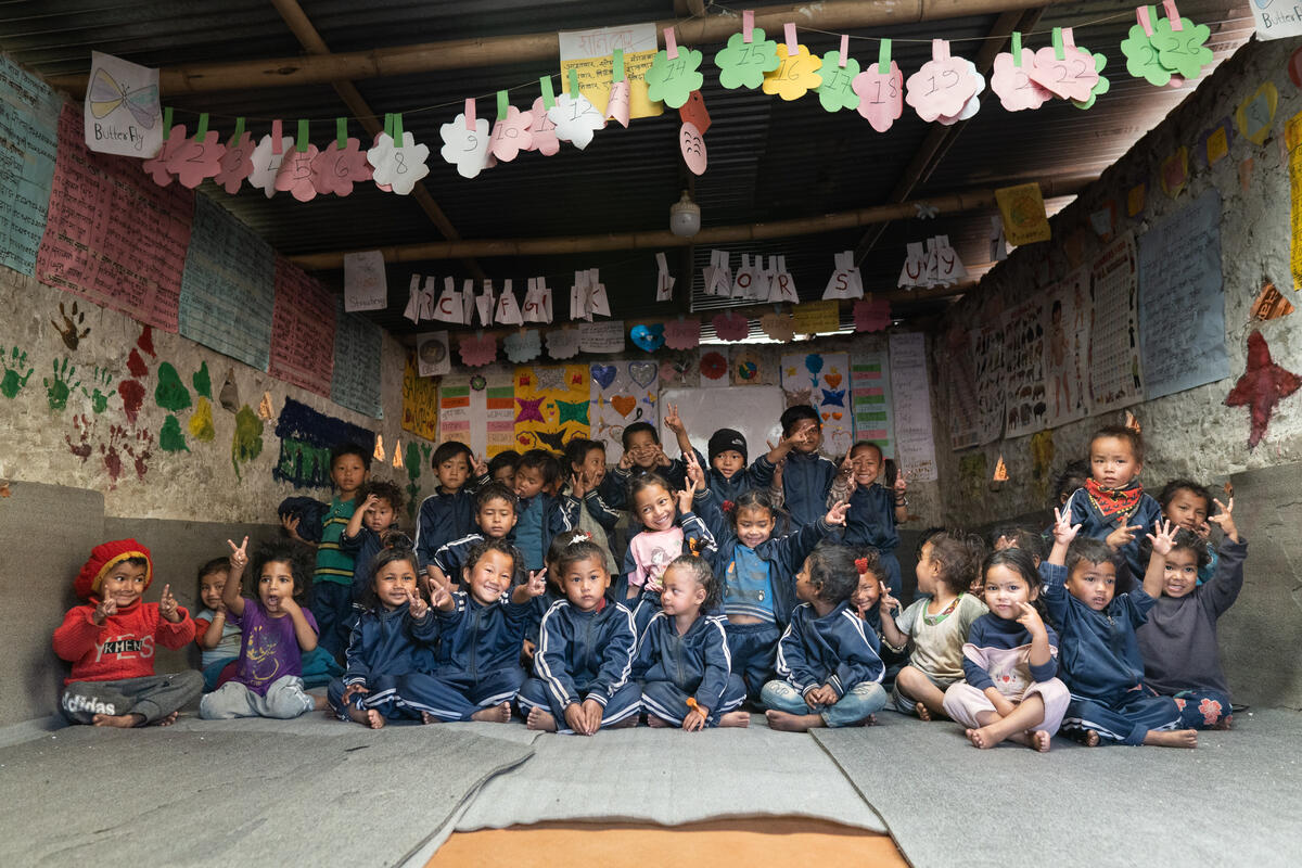 Children in Nepal seated on the floor in a classroom smile at the camera, some making peace signs with their hands.