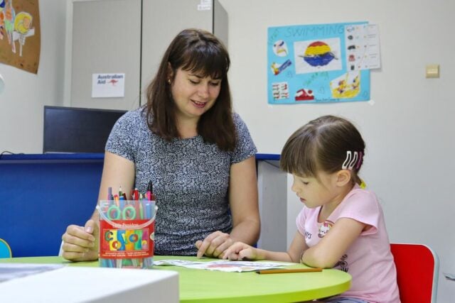 A woman and a young girl sit at a table, focusing on a book together, with colorful markers nearby.