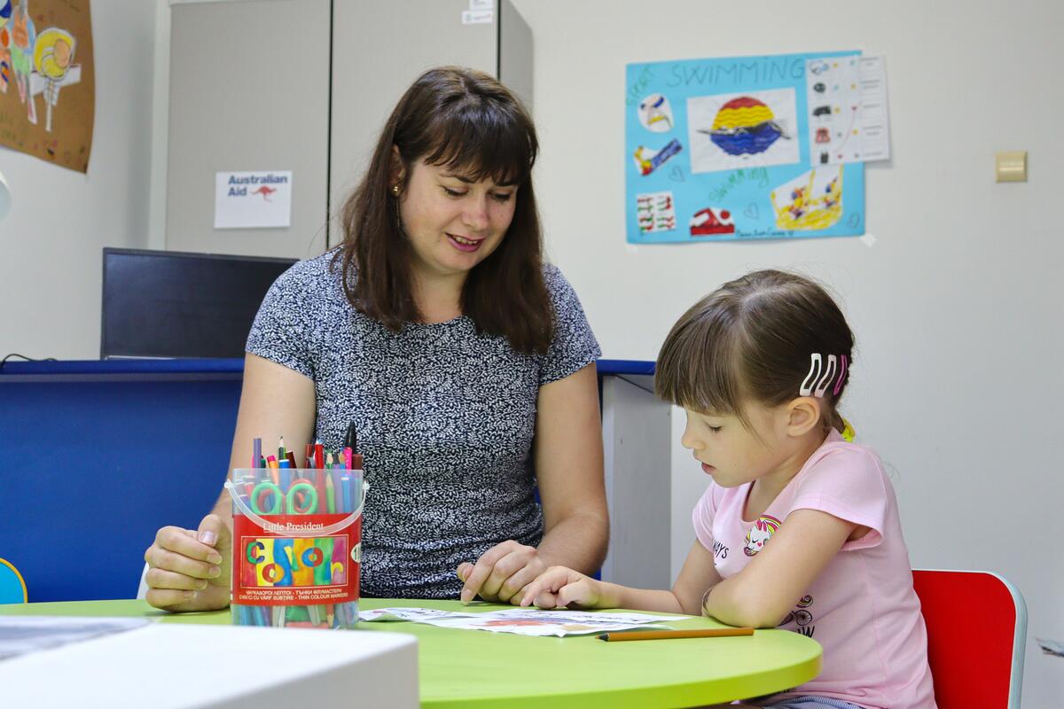 A woman and a young girl sit at a table, focusing on a book together, with colorful markers nearby.