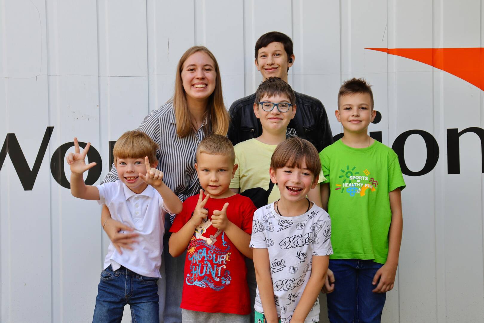 A woman and a group of six children stand in front of a wall painted with a World Vision logo.