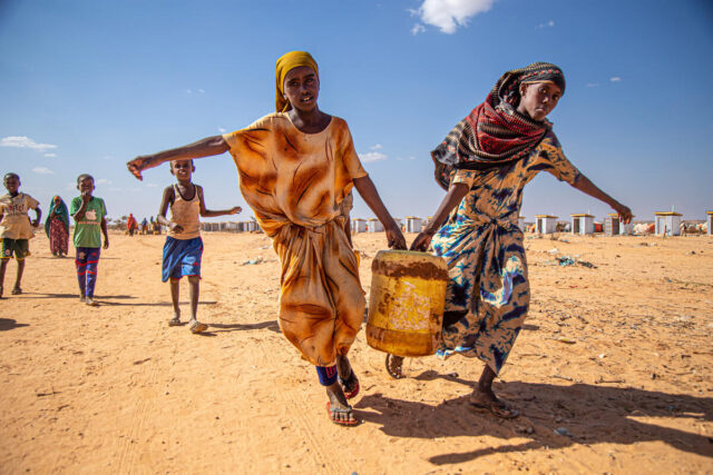 Two girls balance the weight of a yellow jug between them. Their inner hands grip the container with their outer arms extended.
