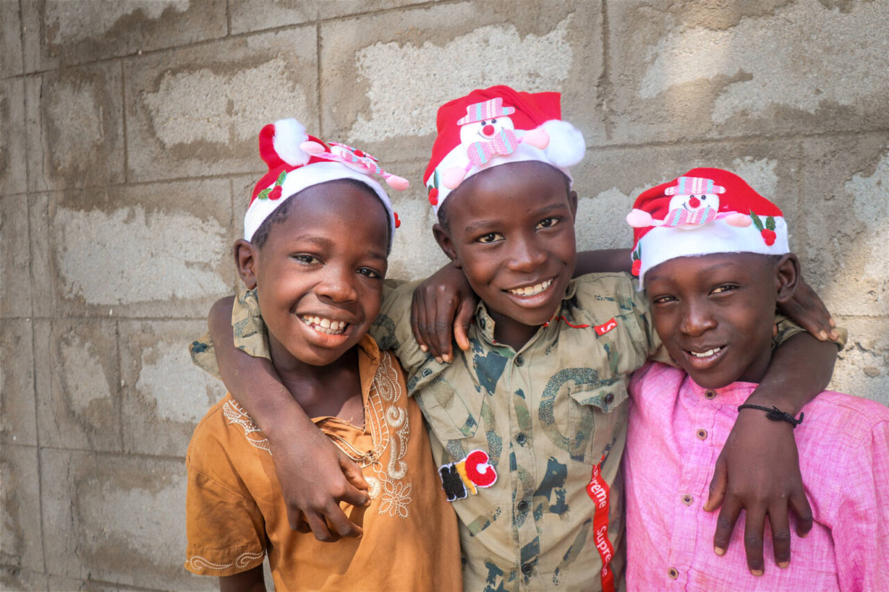 Three boys wearing Santa hats smile directly into the camera, their arms looped around each other&rsquo;s necks.