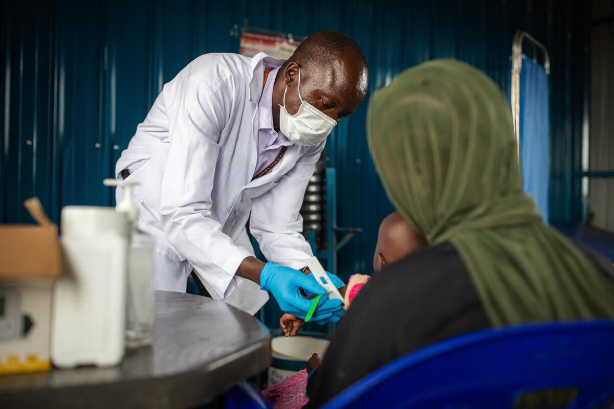 A man in a face mask and a white coat assesses the level of malnutrition in a child held by a woman wearing an olive-colored headscarf.