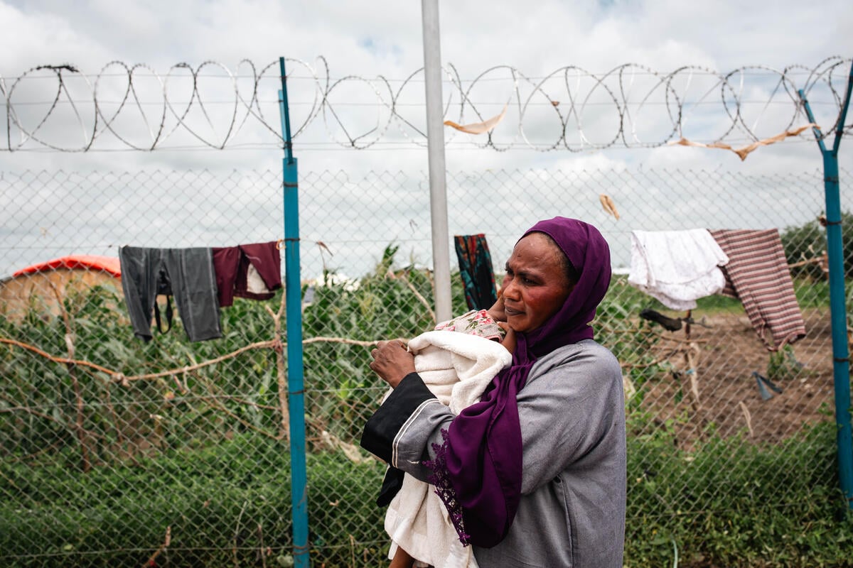 A Sudanese woman in a purple head covering carries a baby in her arms, walking past a barbed wire fence with grass, shrubs, and clothes hanging on a line behind her. 