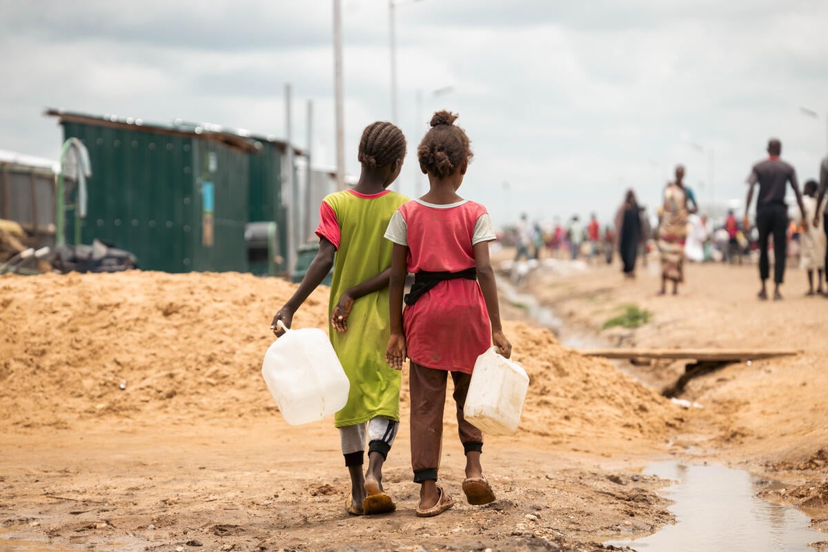 Two girls, each carrying plastic water jugs, walk away from the camera. A stagnate water stream is to their right, while blurred figures can be seen before them. 