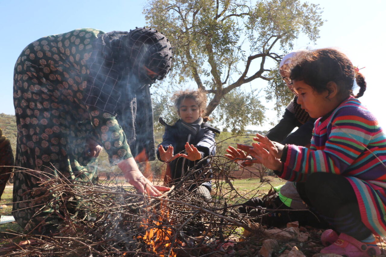 A mother bends down to press her hand on a pile of sticks and stoke a fire to ward off winter&rsquo;s cold from her young daughters.