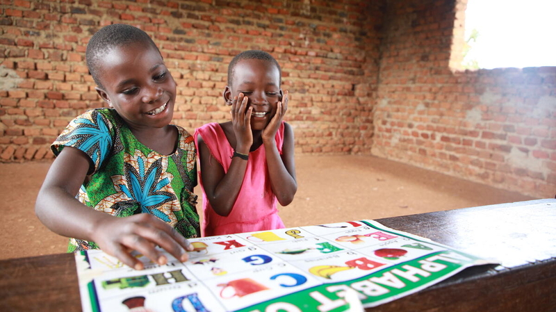 Two Ugandan girls wearing brightly colored shirts smile and laugh as they look at a paper with alphabet letters on it.