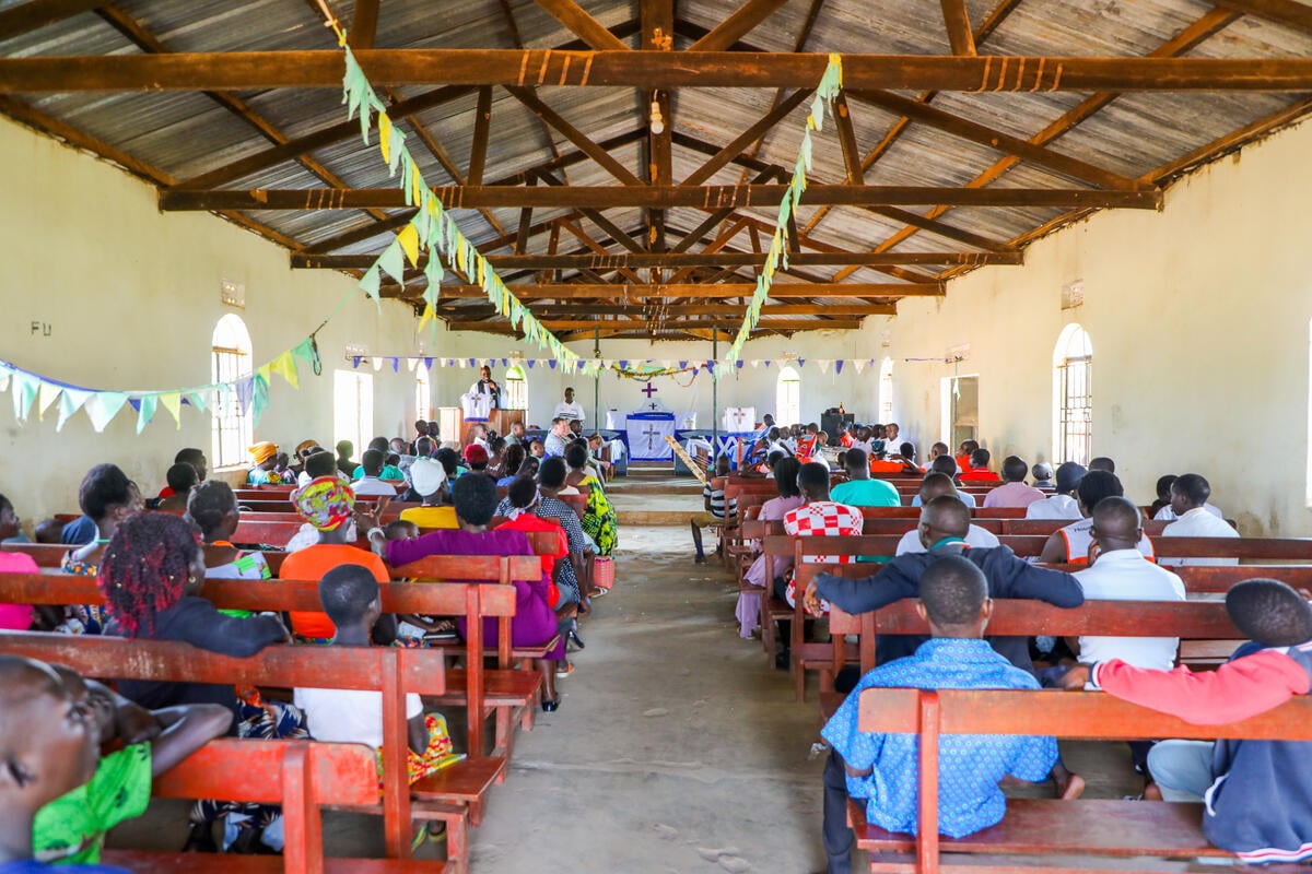 People sit on wooden benches inside a decorated church, listening to speakers at the front near an altar and cross.
