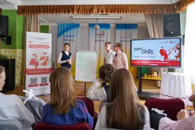 Three young people stand in front of a room filled with adolescents, next to a whiteboard. A screen and banners show drawings and the words &ldquo;Soft Skills,&rdquo; in addition to the World Vision and Responsible Citizens logos.