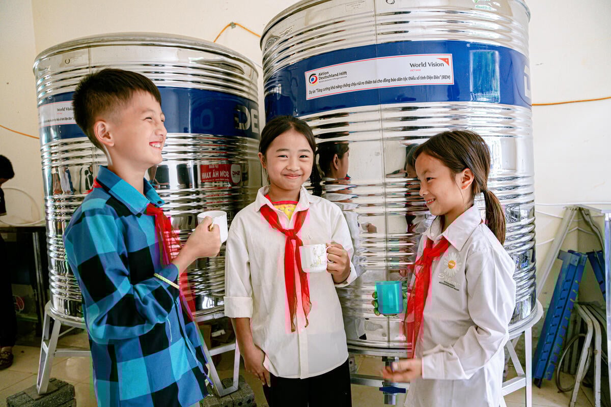 Three children wearing red scarves tied around their necks smile while holding cups, with two large water tanks behind them.