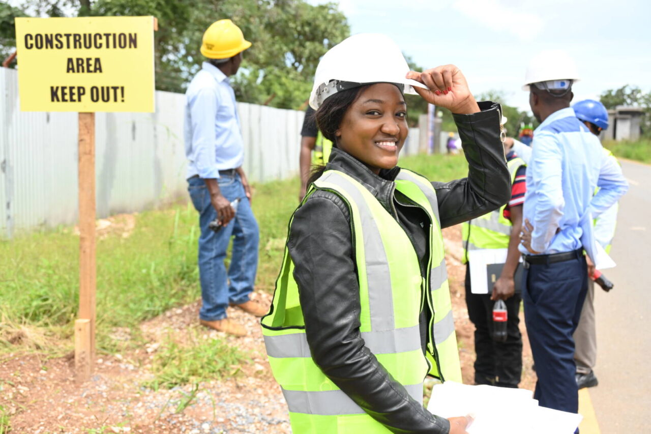 A woman smiles while confidently holding the brim of her hardhat at a construction zone. 