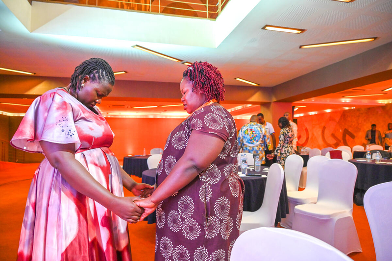 Two women wearing dresses stand holding hands while other men and women stand holding hands in the background, all praying.