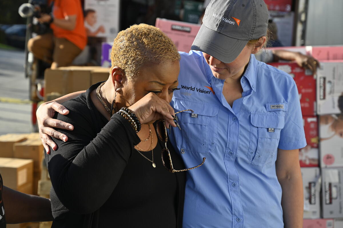 A woman in a World Vision shirt and hat stands with her arm around a woman who wipes her eyes.