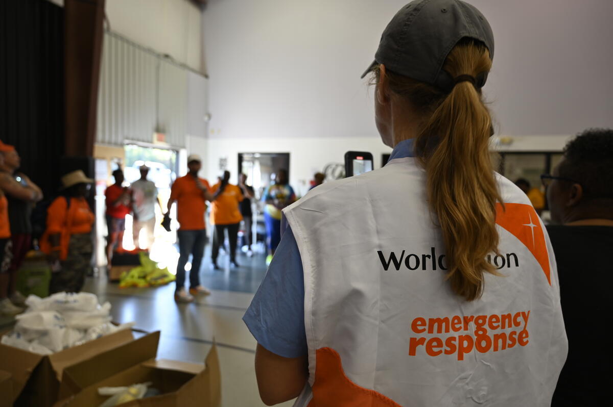 A woman wearing a World Vision vest with a long ponytail stands with her back to the camera, facing a group gathered inside a gym. 