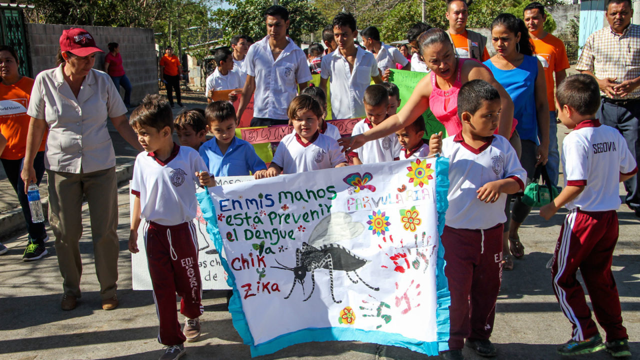 On a sunny day, kids hold a colorful sign with a large mosquito amid a crowd, promoting awareness to prevent the spread of Zika.